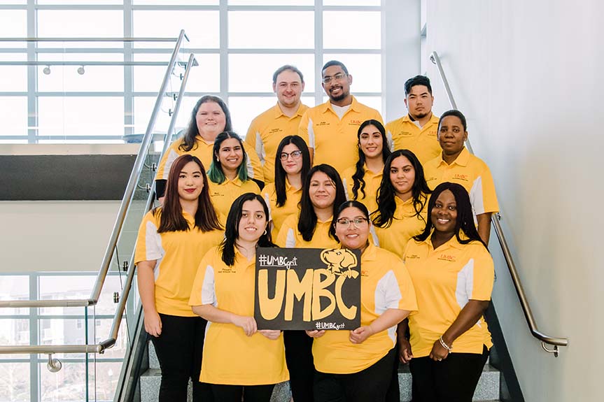 Group of people standing on stairway wearing matching gold shirts and holding a UMBC sign.